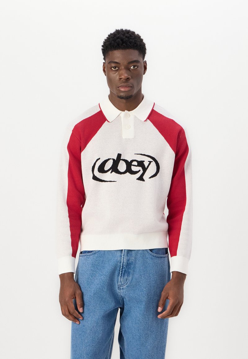 Vit och röd stickad polo tröja med långa ärmar, med en svart "obey"-logotyp i mitten och ribbade cuffs och nederkant. Bärs med blå jeans.