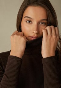 Hnědý žebrovaný rolák s dlouhými rukávy, který má vysoký límec. Modelka drží límec blízko obličeje, čímž ukazuje strukturu látky.