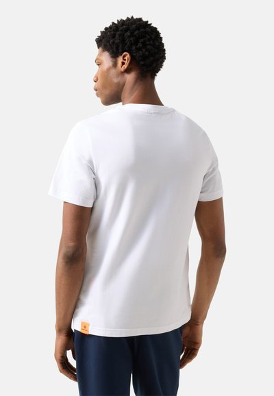 Homme aux cheveux courts et bouclés portant un t-shirt blanc uni à manches courtes et un pantalon bleu foncé, debout dos à nous sur un fond uni.