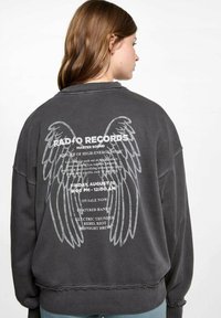 Tmavošedý oversized sweatshir s okrúhlym výstrihom. Na chrbte sa nachádza grafika bielych anjelských krídiel a text udalosti. Materiál vyzerá mäkko.