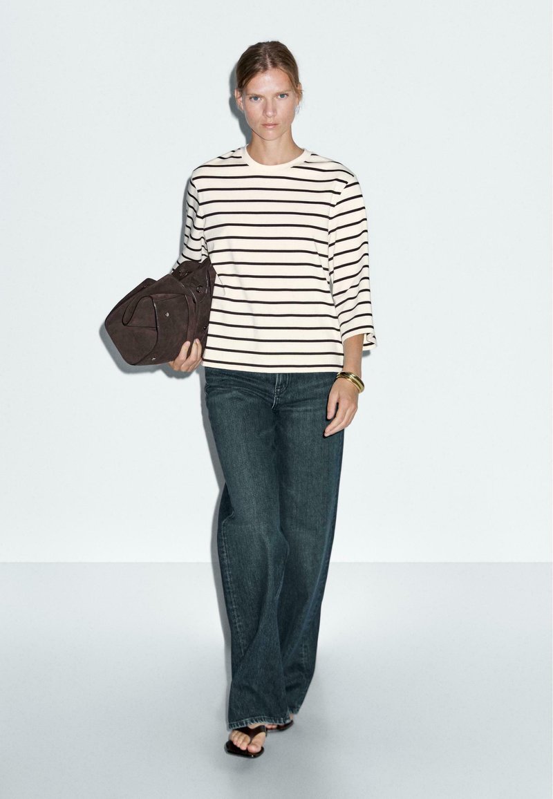 Gestreiftes Langarmoberteil in Creme und Marineblau, kombiniert mit weiten, dunklen Jeans. Das Model trägt eine braune Tasche und schwarze Sandalen.