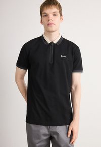 Juodas polo marškinėliai su trumpo užtrauktuko, kreminiu ir pilku apykaklės akcentu, bei nedideliu baltu "BOSS" logotipu ant krūtinės. Medvilnės audinys, trumpomis rankovėmis.