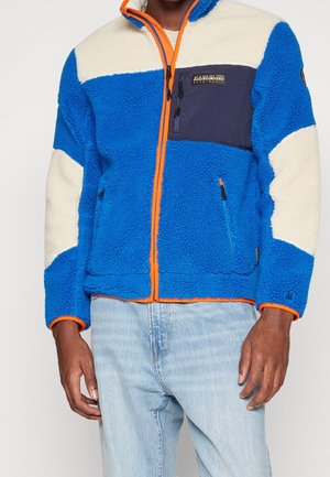 Mann trägt eine blau-weiße Fleecejacke mit orangen Reißverschluss-Akzenten und hellblaue Jeans, Hände locker an den Seiten.