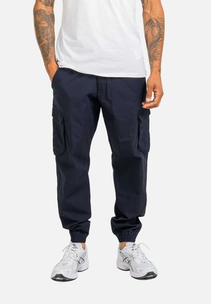 Mann trägt marineblaue Cargohosen mit elastischen Abschlüssen, weiße Sneakers und ein weißes T-Shirt, steht mit einer Hand in der Tasche.