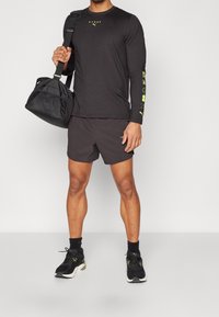 Černá košile s dlouhým rukávem a žlutým logem, doplněná černými šortkami. Černá taška s hladkou texturou. Sportovní boty v černé a bílé barvě.