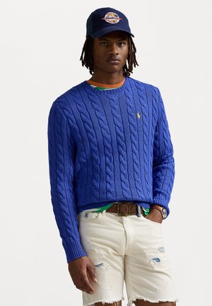 Blauwe kabelgebreide trui met een ronde hals, gecombineerd met versleten witte shorts en een bruine riem. Vergezeld van een marineblauwe pet.