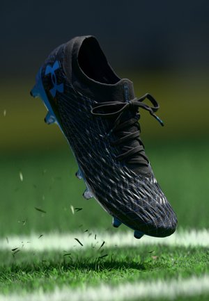 Botas de fútbol negra y azul de Under Armour en el aire sobre un campo de hierba con una línea blanca, levantando pequeños restos de hierba.