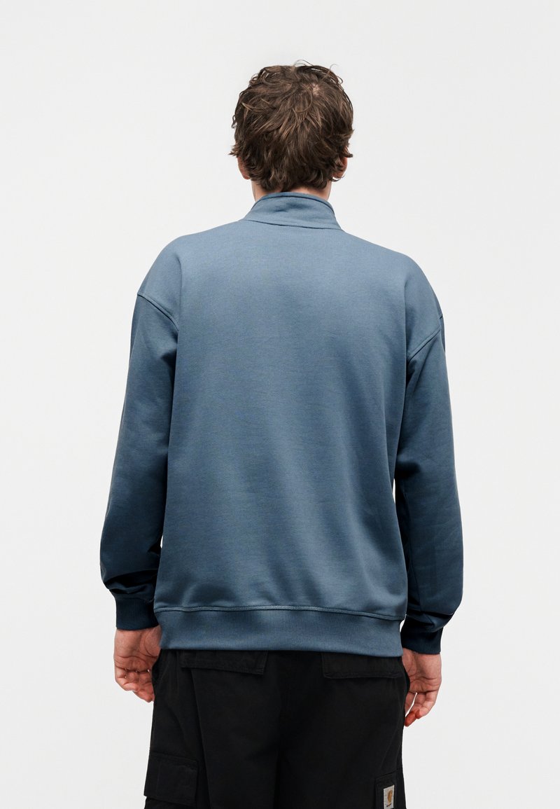 Blue sweatshirt with a high collar, ribbed cuffs and hem, shown from the back. Soft texture and relaxed fit, paired with black cargo pants.