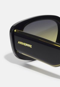 Black sunglasses with a glossy finish and green-tinted lenses. Gold "ARNETTE" branding on the side, shape is angular with thick frames.