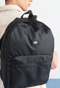 Mochila preta com um tecido liso e durável. Inclui um bolso frontal com zíper proeminente e uma pega na parte de cima. Apresenta o logotipo da marca na frente.