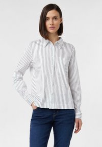White button-up shirt with black vertical stripes, long sleeves, pointed collar, and a pocket on the left chest; paired with dark jeans.