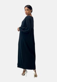 Abito lungo nero a maniche lunghe con tessuto texturizzato, vestibilità ampia e arricciatura in vita. Indossato con scarpe con tacco alto bianche.
