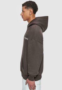 Brauner Hoodie mit vorderer Tasche, Kapuze mit Kordelzug und gerippten Bündchen. Verfügt über Grafikttext auf der linken Brust und ein unregelmäßiges Design auf der Rückseite.