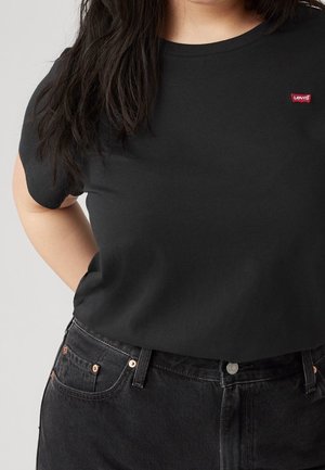 Personne portant un t-shirt Levi's noir et un jean noir, avec de longs cheveux foncés sur l'épaule, sur un fond clair uni.