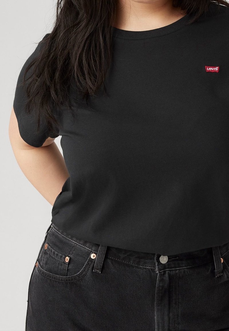 Personne portant un t-shirt Levi's noir et un jean noir, avec de longs cheveux foncés sur l'épaule, sur un fond clair uni.