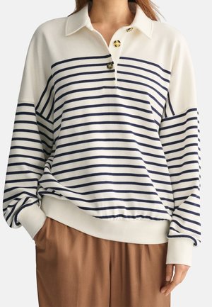 Kobieta ubrana w białą bluzkę z długim rękawem z poziomymi granatowymi paskami, guziki w kolorze żółwia oraz jasnobrązowe spodnie, z ręką w kieszeni.