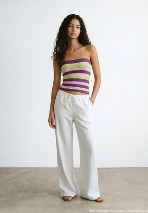 Femme aux longs cheveux bouclés portant un top tube rayé et un pantalon blanc ample à cordon, debout sur un sol gris contre un mur blanc.