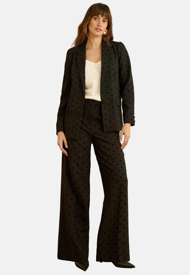 Costum negru cu buline, format dintr-un sacou croi ajustat și pantaloni largi. Prezintă o textură fină și o bluză contrastantă, de culoare deschisă, dedesubt.