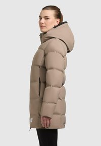 Beige puffer jacket with a hood, quilted design, front zip closure, side pockets, and a logo tag near the hem. Smooth texture.