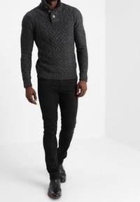 Pull en tricot gris foncé avec un motif texturé, col rabattable et bouton en bois. Porté avec un jean noir et des bottines.
