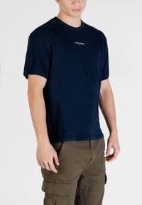 T-shirt in cotone blu navy, collo rotondo, maniche corte, con un piccolo logo bianco "TOMMY JEANS" sul petto, indossata con pantaloni cargo.