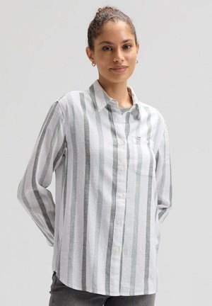 Kobieta z włosami związanymi w koka, nosząca złote kolczyki-koła oraz białą koszulę w pionowe szare paski, stojąca na tle jednolitego jasnego tła.