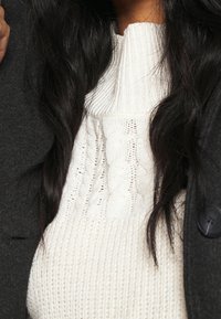 Suéter de punto cable beige con cuello alto, combinado con un abrigo gris oscuro. El suéter presenta patrones texturizados y detalles acanalados.