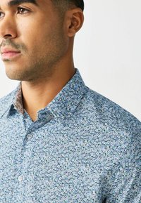 Ziedu raksta krekls gaiši zils, ar mazām, daudzkrāsainām ziediem. Pogājamais apkakle, teksturēts audums, izteikta kontrastējoša iekšējā apkakle.