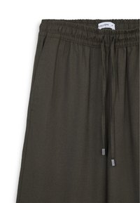 Dunkel olivgrüne Hose mit elastischem Bund und Kordelzug, mit glatter Textur und silberfarbenen Hardware-Details. Markenetikett sichtbar.