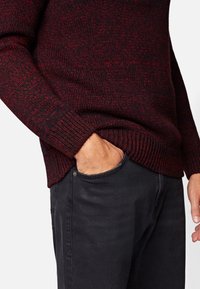 Pull en tricot rouge foncé et noir, avec un motif texturé et des poignets côtelés. Associé à un jean gris foncé, main dans la poche.