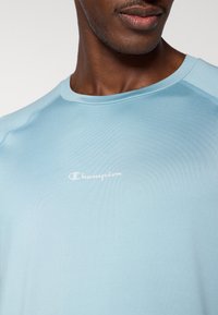 Αθλητική μπλούζα σε ανοιχτό γαλάζιο χρώμα, κατασκευασμένη από απαλό ύφασμα που απομακρύνει την υγρασία. Διαθέτει στ ρογγυλή λαιμόκοψη και λευκό λογότυπο "Champion" στο στήθος.