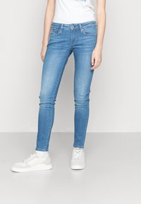 Person klädd i ljusblå slim fit-jeans, vita sneakers och en vit t-shirt som står mot en enfärgad ljus bakgrund.