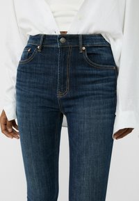 Jeans de denim azul oscuro de cintura alta con costuras en color beige, cinco bolsillos y cierre de botón metálico. Tejido suave y texturizado.