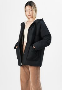 Ung kvinne i svart polstret jakke med fleecekrage, beige crop top og brune cordbukser, som står foran en enkel hvit bakgrunn.