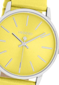 Montre ronde avec un cadran mat jaune vibrant, index et aiguilles argentés. Elle possède un bracelet en cuir jaune texturé et un boîtier en acier inoxydable.