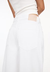 Weiße Denim-Hosen besitzen eine hohe Taille, weit geschnittene Beine und Gesäßtaschen, mit einem hellbraunen Lederpatch am Bund.