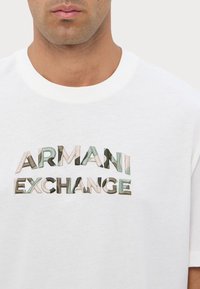 T-shirt in cotone bianco con il logo "ARMANI EXCHANGE" in ricamo testurizzato multicolore sul petto. Design classico con collo a giro.