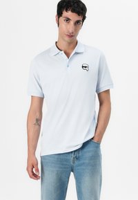 Lyseblå polo-skjorte i bomull, med et svart og hvitt grafisk logo på brystet. Korte ermer og krage.