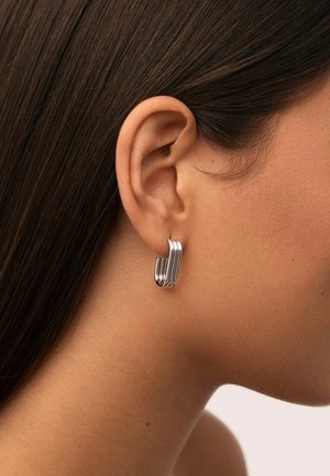 Orecchino a cerchio in argento, caratterizzato da un design stratificato e curvato con una finitura liscia. Indossato all'orecchio sinistro con capelli castani scuri sullo sfondo.
