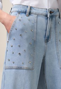 Jeans de mezclilla azul claro con grandes bolsillos delanteros adornados con brillantes cristales dispersos y detalles de costura en contraste.