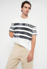 Λευκό βαμβακερό t-shirt με ναυτικό οριζόντιες ρίγες, που διαθέτει την επωνυμία "GUESS" στο ύψος του στήθους σε μαύρο χρώμα. Χαλαρή γραμμή και στρογγυλή λαιμόκοψη.