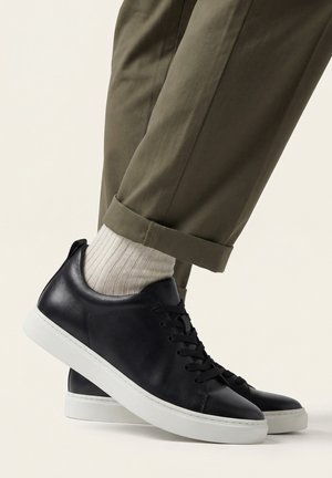 Osoba w oliwkowozielonych spodniach z mankietami, białych prążkowanych skarpetkach i czarnych skórzanych trampkach z białymi podeszwami, stojąca na jasnym tle.