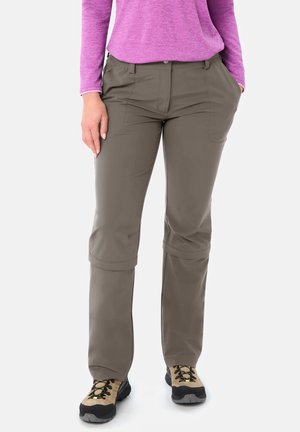 Vrouw die bruine om te zetten wandelbroeken draagt, tan- en zwarte wandelschoenen en een paarse lange mouwen shirt, staand met één hand in de zak.