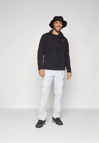 Fekete gyapjú cipzáras dzseki, fekete póló, világosszürke zsebes nadrág cipzárakkal, fekete vödörkalap és fekete túracipő. Álló póz.