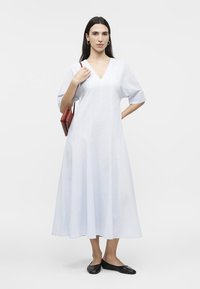 Γυναίκα που φοράει ένα φαρδύ λευκό φόρεμα μέσης μήκους με V λαιμόκοψη, μαύρα χαμηλά παπούτσια, χρυσούς κρίκους στα αυτιά και κρατάει μια μικρή κόκκινη τσάντα στον ώμο της.