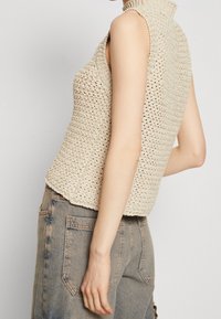 Beige gestricktes ärmelloses Oberteil mit hohem Kragen; verfügt über ein strukturiertes Muster und eine figurbetonte Form. Kombiniert mit hellblauen Jeans.