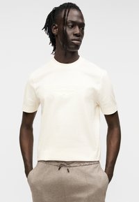 Κρεμ κοντομάνικη μπλούζα με ανάγλυφο λογότυπο Aston Martin στο μπροστινό μέρος, κατασκευασμένη από απαλό βαμβακερό ύφασμα.