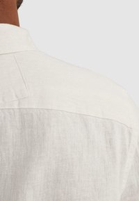 Κοντινό πλάνο στο πάνω μέρος της πλάτης και στον ώμο ενός ατόμου με ανοιχτό μπεζ λινό πουκάμισο με γιακά και ραφές.