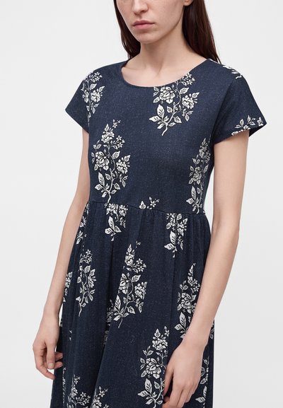 Femme portant une robe bleu marine avec des motifs floraux blancs, des manches courtes et un col rond.