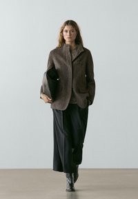 Brown textured blazer with a stand collar, two front pockets, worn over a gray top, paired with wide black trousers and ankle boots.